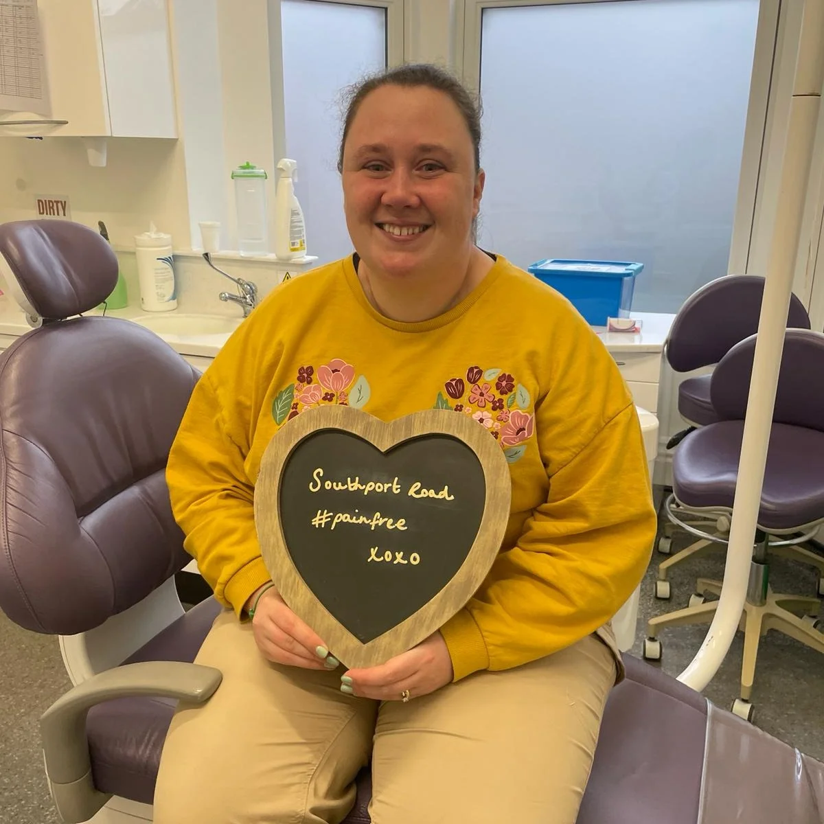 A patient in the dental chair holding a heart-shaped board reading "Southport Road #painfree"