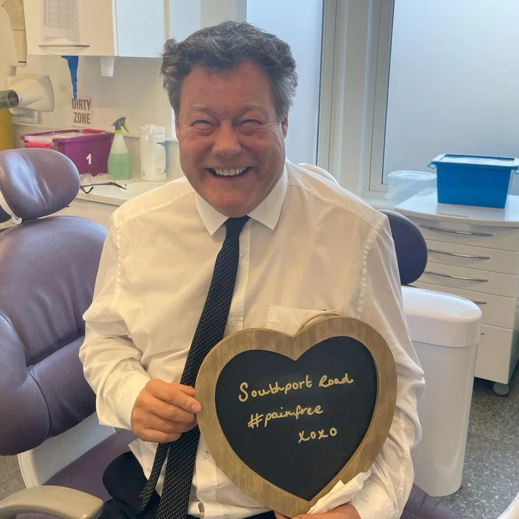 A patient in the dental chair holding a heart-shaped board reading "Southport Road #painfree"
