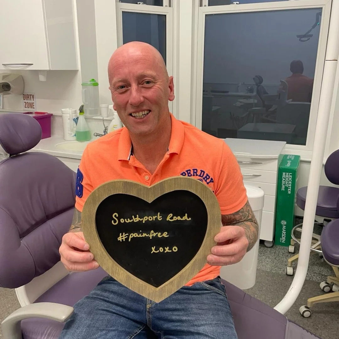A patient in the dental chair holding a heart-shaped board reading "Southport Road #painfree"